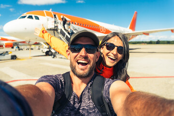 Cheerful caucasian couple taking selfie in front of passenger airplane before boarding flight symbolizing travel, tourism, adventure, and vacation lifestyle.