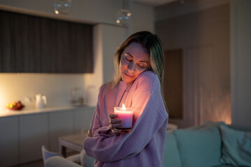 Happy peaceful middle aged woman standing alone indoor holding glowing candle enjoying calm evening warmth. Emotional female smiling feeling pleasure in deep gratitude at comfort home. Thank you life.