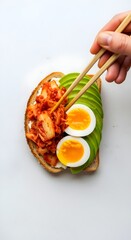 Mindful preparation of a modern avocado toast, with a hand using chopsticks to add probiotic kimchi and egg, creating a functional food meal for gut health and mood support.

