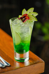 Green apple cocktail served in a tall glass with ice, green syrup, apple slices, and an edible flower garnish.