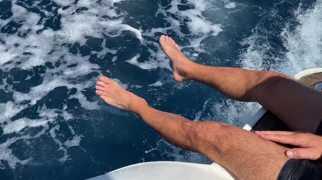High view shot of unrecognisable man sitting on aft deck of sailing motorboat with legs put inside the water, refreshing riding on speed boat on the sea with amazing blue water, exploring summer