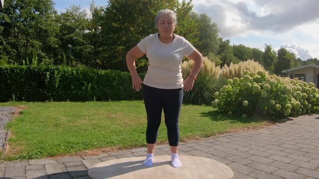 Overweight adult woman doing a standing forward bend to touch ground in garden, strengthening back for flexibility. Home workout plan for seniors seeking a healthy lifestyle