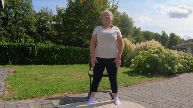 Front view of overweight adult woman doing squats on chair in garden, strengthening legs and knees to improve flexibility. Home workout plan for seniors seeking a healthy lifestyle