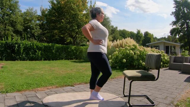 Overweight senior woman lifting legs with help of chair in garden, strengthening core and improving flexibility. Outdoor home workout plan for seniors seeking a healthy lifestyle