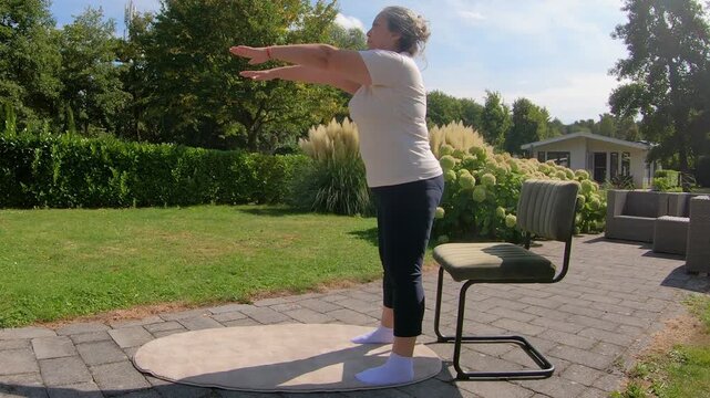 Overweight adult woman doing squats with arm stretches on chair in garden, strengthening legs and knees to improve flexibility. Home workout plan for seniors seeking a healthy lifestyle