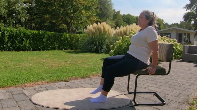 Overweight adult woman doing sit-ups while sitting on chair in garden, strengthening core by raising and lowering legs. Outdoor home workout plan for seniors seeking a healthy lifestyle