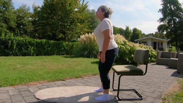 Overweight adult woman doing squats on a chair in the garden, strengthening legs and knees to improve flexibility, outdoor home workout plan for seniors seeking a healthy lifestyle