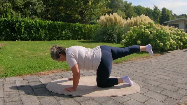 Overweight senior woman practicing back kicks on garden floor, strengthening buttocks and legs to improve flexibility. Home workout plan for seniors seeking a healthy lifestyle