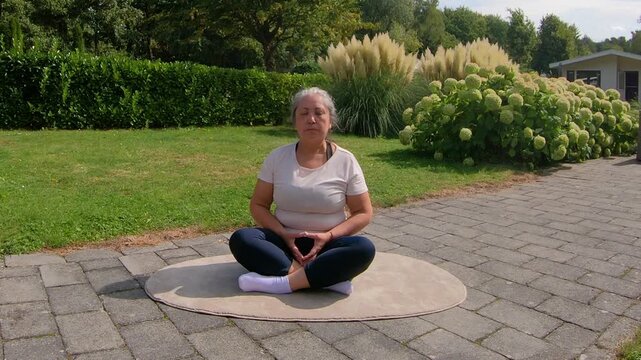 Overweight adult woman doing some relaxing breathing sitting on garden floor, warming up or relaxing after exercise. Home workout plan for seniors seeking a healthy lifestyle