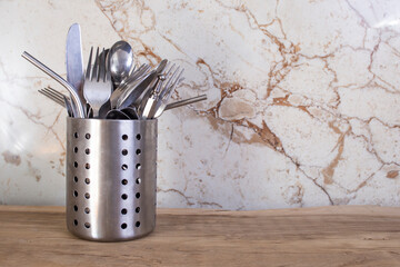 Spoons, forks and stainless steel straws in a strainer container , on wood table with beige marble backsplash, abstract kitchenware backdrop