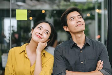 Happy Asian man and woman looking up with positive expressions in office. Creative team standing in front of glass wall with sticky note, concept of inspiration, teamwork and planning.