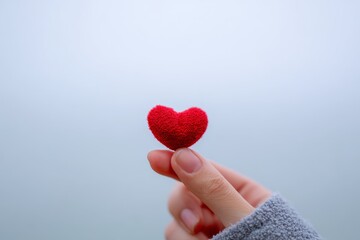 Fototapeta premium A small, textured red heart is held gently by a hand, showcasing a simple yet heartfelt gesture against a soft, light gray backdrop.