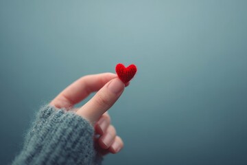 Fototapeta premium A delicate red heart-shaped object rests gently on a finger, held by a hand wearing a gray knit sleeve against a soft, muted backdrop.