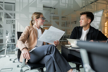 Business meeting between two professionals discussing documents in a modern office environment with glass walls and a collaborative atmosphere