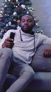 Young handsome African man in casual clothes sitting on sofa and viewing television. Disgruntled guy switching TV channels. Indoors.