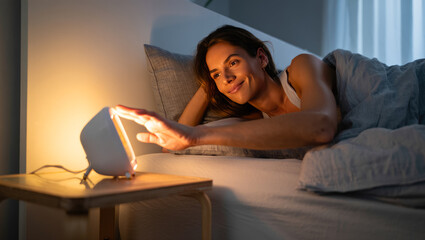 Fototapeta premium Woman wakes up with the help of a smart alarm clock with sunrise simulation in a cozy bedroom. Modern wake up light technology for healthy sleep, natural morning routine, and stress-free lifestyle.