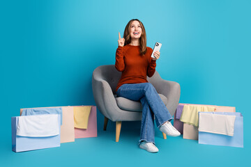Smiling young woman sitting on chair amid shopping bags, holding smartphone in vibrant blue background