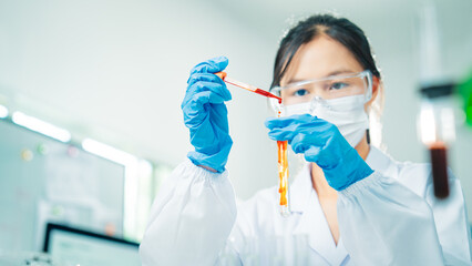Female scientist wearing protective gloves and mask transferring red liquid into a test tube,...
