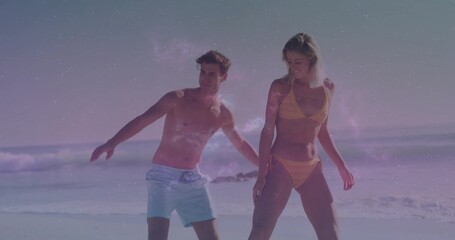 Walking couple wearing yellow bikini and light blue swim trunks at sandy beach, with ocean waves