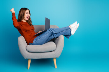 Excited young woman sitting on a chair holding a laptop against a blue background, celebrating...