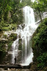Obraz premium A person standing at the base of a majestic waterfall, completely dwarfed by its powerful cascade. 