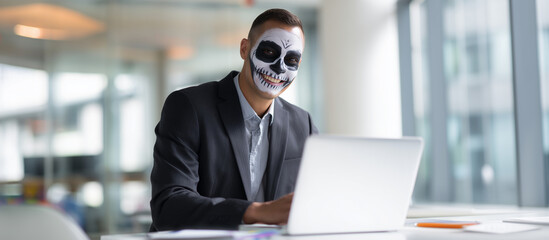 Hispanic businessman with skull face paint smiling while working on laptop in modern office environment.