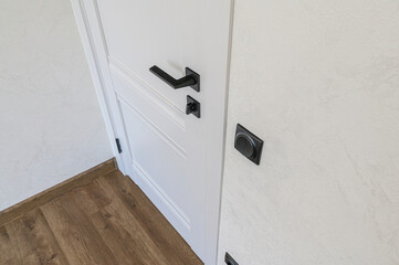Close-up of a white door with a sleek black handle, paired with a black switch against a textured cream wall. Wooden floor trim visible