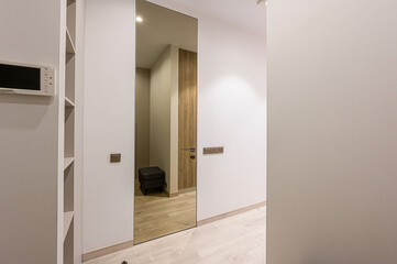 sleek hallway with a full-length mirror reflecting a room. The walls are white with a light wooden floor. An intercom and shelves adorn one side