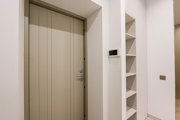 Neutral toned door in a white hallway. A shelving unit and security monitor are adjacent