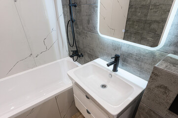 Modern bathroom with white tub and sink, black fixtures, marble accents, and a lit mirror. Clean, stylish design