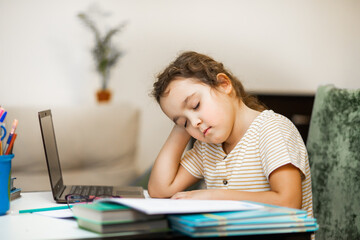 Little cute girl pupil at home making homework. Kid sleeping resting head on books. Child studying online on laptop. Kid education.