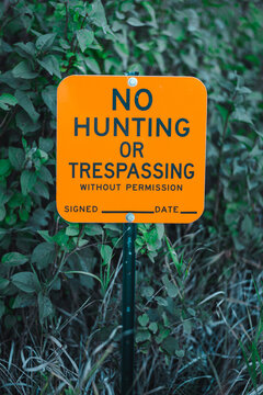Neon orange reflective &ldquo;No Hunting or Trespassing Without Permission&rdquo; sign on post, posted warning in dense green foliage, outdoor safety notice in rural woods, forest, or private property boundary