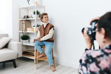 Bright living space portrait featuring a young model seated on a wooden stool while a photographer captures a candid moment with warm, natural light.