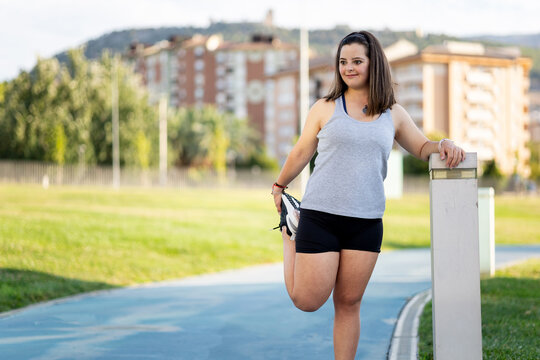 A girl between 20 and 30 years old with Down syndrome is doing sports outdoors.The girl is stretching her leg back. Concept for people with trisomy 21.