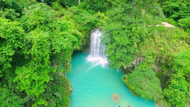Aerial view of a turquoise waterfall cascading into natural pools with people swimming, surrounded by lush tropical jungle, Kawasan Falls, Philippines.