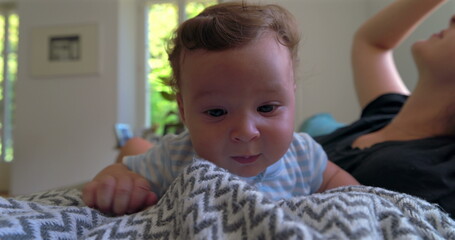 Fototapeta premium Baby lying on a blanket, looking slightly frustrated and emotional, in a warm home setting with natural light, while mother observes in the background