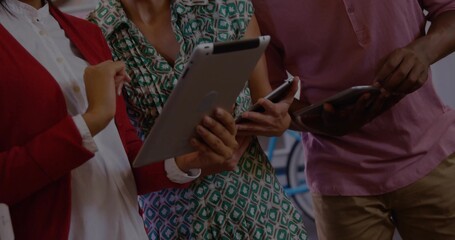 Standing three coworkers wearing casual attire collaborating in office, with tablets and smartphone