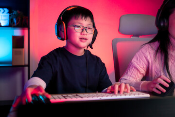 Two happy siblings wearing headphones and playing on a desktop computer in a colorful gaming setup
