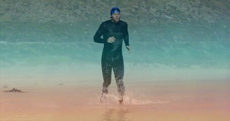 Athlete in black wetsuit running onto rocky lakeshore from shallow water, with swim cap and goggles