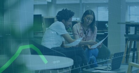 Analyzing printed charts, two coworkers leaning forward in office lounge with laptop and smartphone