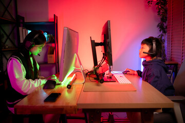 Two happy siblings wearing headphones and playing on a desktop computer in a colorful gaming setup © Wosunan