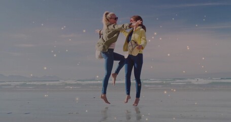 Leaping friends wearing olive and yellow jackets on beach near water's edge, with crossbody purses
