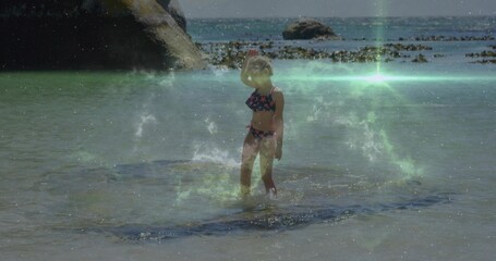 Obraz premium Wading female child in floral swimsuit moving through ocean at rocky shore, with rocks and seaweed