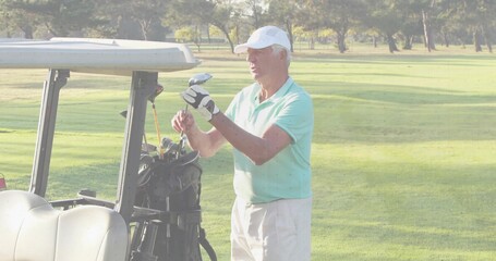 Senior male golfer wearing polo and cap selecting club from bag beside cart on fairway