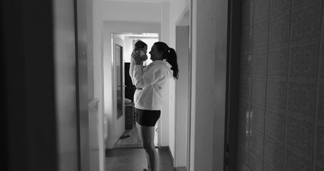 Mother lifting her baby in a dimly lit hallway, sharing a joyful and tender moment. The black and...