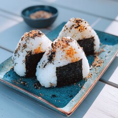 Three triangular onigiri, rice balls, topped with seaweed and sesame seeds, sit on a teal-colored rectangular plate. A small bowl of seasoning is visible in the background