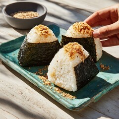 Three triangular onigiri, topped with sesame seeds, on a teal plate. A hand reaches for one