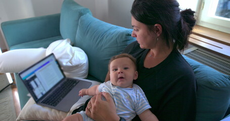 Mother working on laptop while holding baby at home, balancing between work and parenting, multitasking in a cozy living room environment