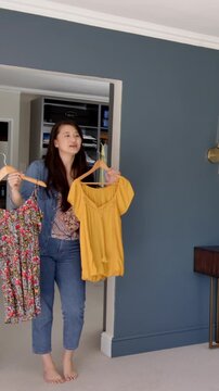 Woman holding two dresses on hangers, standing barefoot between closet and teal painted wall