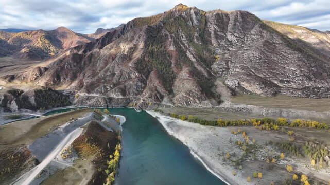 An aerial panoramic view of a wide, turquoise river flowing through a mountain valley with rocky, rugged terrain and scattered golden autumn trees